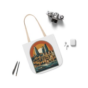 May include: A white tote bag featuring a colorful graphic illustration of a coastal city skyline with tall flame-shaped skyscrapers, a sunset, and boats on the water. The design is enclosed in an arched frame with warm orange and blue tones.
