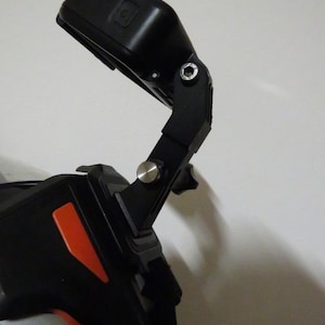 Puede incluir: Una cámara de acción negra montada en un brazo ajustable negro sujeto a un casco. El soporte cuenta con un tornillo de pivote de metal plateado y una base de plástico negro con una pieza de acento de color naranja brillante en el casco.