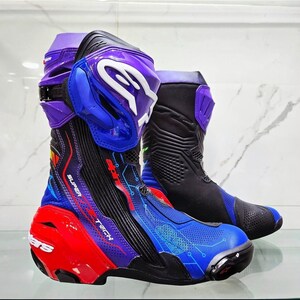 Puede incluir: Un par de botas de carreras para motocicleta Alpinestars Supertech con un esquema de colores vibrantes en morado, azul y rojo. Las botas incluyen un logotipo blanco prominente en la espinillera, paneles negros texturizados y gráficos de circuitos.