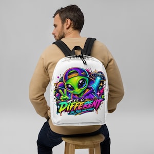 Può includere: Uno zaino bianco con una grafica colorata di un alieno verde che indossa un berretto e una felpa viola, mentre fa il segno della pace. Il design include un UFO, pianeti e il testo "DIFFERENT BE YOUR OWN PLANET" in vivaci colori neon.