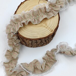 May include: A length of ruffled fabric trim featuring a light green and pink checkered pattern, accented with a delicate white lace border along the edges. The trim is draped over a circular wooden slice with visible bark on the outer edge.