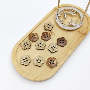May include: A collection of ten flower-shaped buttons made from brown coconut shell, each featuring four central holes and etched petal details. The buttons are arranged on an oval bamboo tray next to a silver-toned metal stand with bamboo motifs.