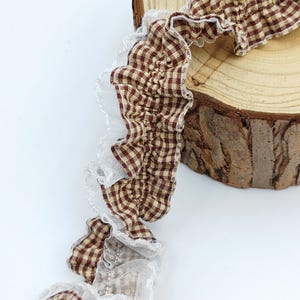 May include: A brown and white gingham fabric trim featuring a ruffled edge with a delicate white sheer lace border. The fabric strip is gathered and textured, showcasing a classic checkered pattern throughout the length of the material.