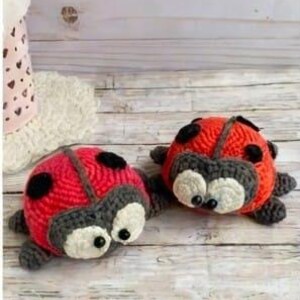 Puede incluir: Dos peluches de mariquita tejidos a ganchillo con cuerpos de color rojo brillante con puntos negros, cabezas de color gris oscuro y ojos grandes de color blanco con pupilas negras. Tienen patas pequeñas y una línea gris oscuro en el dorso.