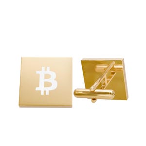 May include: A pair of square gold-toned cufflinks featuring a white Bitcoin symbol centered on the front face. The cufflinks have a polished metallic finish and a standard hinged toggle mechanism on the back for securing through shirt cuffs.