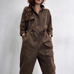 May include: A dark brown long-sleeve jumpsuit featuring a button-down front, a collared neckline, and a drawstring waist. The garment includes a chest pocket with a flap and a large cargo-style pocket on the left leg, finished with cuffed hems.