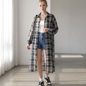 May include: A long-sleeved, button-down shirt featuring a black, white and grey plaid pattern. The shirt is worn open over a plain white vest top and blue denim shorts, paired with black and white trainers and a black shoulder bag.