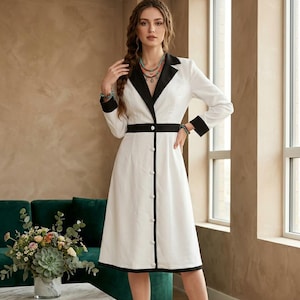 May include: A white midi-length dress featuring a black notched lapel collar, black sleeve cuffs, and a black vertical trim running down the centre. The dress includes a black waist band and a row of white buttons extending from the waist to the hem.