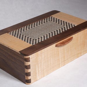 Pode incluir: Uma caixa de madeira com um inlay xadrez na tampa. A caixa é feita de madeira de noz e bordo e tem uma pequena alça de madeira.