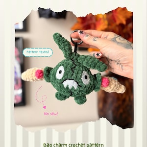 Peut inclure: Une breloque de sac en crochet vert foncé avec un anneau porte-clés noir, dotée d'yeux blancs, d'une bouche blanche et de bras beige clair avec des accents circulaires roses. Le texte inclut TRUBBLE, Pattern tested, No sew! et Bag charm crochet pattern by @kimmis.crochet.
