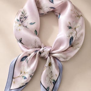 May include: A square scarf in a soft pale pink hue featuring a delicate floral pattern with white blossoms, green leaves, and dark blue accents. The scarf is tied in a knot and finished with a light blue border and a thin black inner trim.