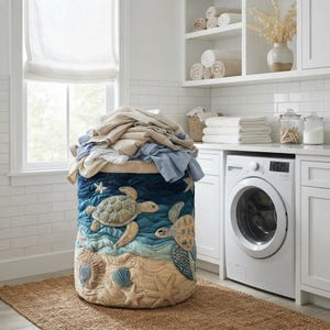 Può includere: Un cesto portabiancheria cilindrico in tessuto con una scena oceanica trapuntata raffigurante tartarughe marine, conchiglie e stelle marine nei toni del blu, verde acqua e beige. Il cesto è riempito con una pila di vestiti piegati nei toni neutri e blu.