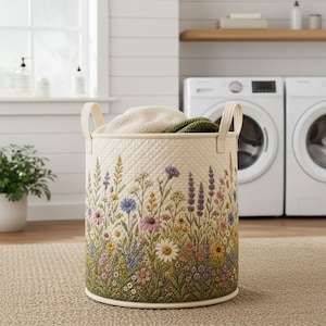 May include: A cylindrical laundry hamper with a cream-coloured quilted exterior featuring a vibrant, multi-coloured botanical print of wildflowers, including daisies, lavender, and cornflowers. The hamper has two vertical fabric handles on the sides.