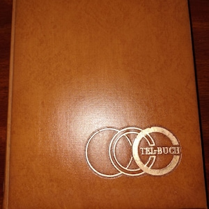 May include: A brown leather-bound book with the word "TEL-BUCH" in gold lettering. The cover features a gold emblem of three interlocking circles. The book is rectangular and appears to be closed.
