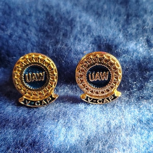 May include: Two gold-colored UAW V-CAP pins. Each pin has a circular design with the letters "UAW" in a blue center. The pins have a decorative border and a banner that reads "V-CAP" in black.
