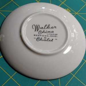 May include: A white ceramic saucer base featuring black printed text that reads Walker China, Vitrified, Bedford, Ohio, and Chalet. The underside of the saucer is smooth and circular, displaying the manufacturer's mark clearly in the center.