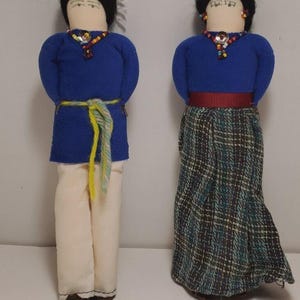 May include: Two handmade dolls dressed in traditional attire. One doll wears a blue top, cream pants, and a yellow and green belt. The other doll has a blue top, a plaid skirt, and a red belt. Both dolls have black hair and beaded necklaces.