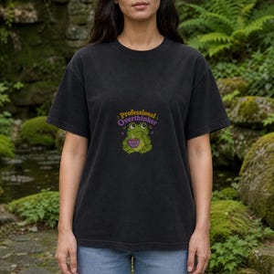 May include: A black short-sleeved t-shirt featuring a graphic of a green frog holding a purple cup with steam rising. Above the frog, the text reads Professional Overthinker in yellow and purple letters. The shirt is paired with light blue jeans.