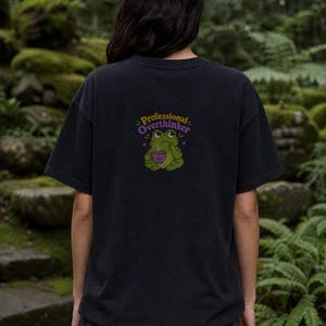 May include: A black short-sleeve t-shirt featuring a graphic on the back of a green frog holding a purple teacup. Above the frog, the text reads Professional Overthinker in yellow and purple letters, accompanied by small purple star accents.