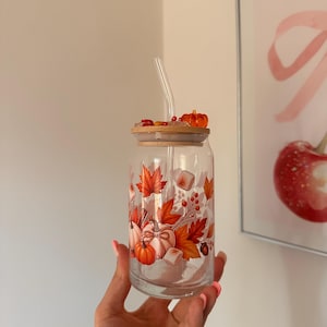 Puede incluir: Un vaso de cristal transparente con un estampado otoñal de calabazas naranjas, hojas de arce rojas, bayas y malvaviscos. El vaso incluye una tapa de bambú con detalles decorativos de resina y una pajita de cristal para beber.