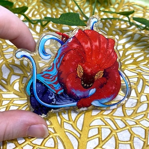 May include: A clear acrylic charm featuring a vibrant red creature with a large, fan-like crest and yellow patterned markings. The design includes swirling blue water elements, a dark purple base, and subtle glitter accents throughout the transparent material.