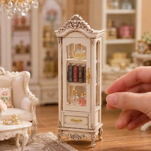 Pode incluir: Uma vitrine em miniatura de madeira em tom branco sujo com detalhes esculpidos, porta de vidro e uma pequena gaveta. As prateleiras internas contêm frascos de perfume e livros em miniatura. Possui ferragens e pés em tom dourado.