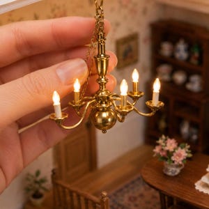 Op de afbeelding: Een miniatuur kroonluchter van goudkleurig metaal met vijf armen, elk voorzien van een kleine witte kaars met een brandend lichtje. Het armatuur hangt aan een fijn gouden kettinkje en is bedoeld voor een poppenhuis of miniatuurweergave.