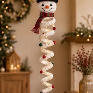 May include: A hanging crochet snowman decoration featuring a white head with a black top hat, a red and green striped scarf, and a long, white, spiral-shaped body adorned with small red and green pom-poms and integrated warm white string lights.