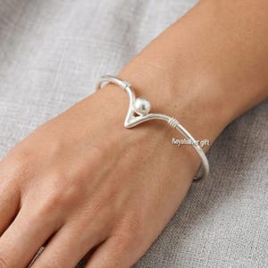 Puede incluir: Una pulsera rígida de tono plateado con un diseño central en forma de V y una cuenta esférica única en el vértice. La banda metálica incluye detalles sutiles de alambre enrollado a ambos lados del punto central, creando una pieza abierta y estructurada.