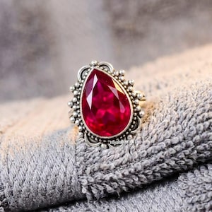May include: A silver ring featuring a large, pear-shaped, faceted deep red gemstone set in a decorative metal frame with small bead-like accents along the outer edge. The text royalsilvergift is visible on the lower portion of the ring setting.