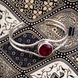 Può includere: Un bracciale rigido in argento con un design a doppia fascia che converge al centro per sostenere una singola pietra preziosa rotonda di colore rosso intenso. Il metallo ha una finitura lucida e il testo royalsilvergift è visibile vicino.