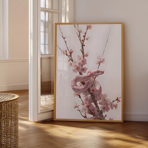May include: A framed vertical wall art print featuring a pale pink snake coiled around a slender branch covered in delicate light pink cherry blossoms. The snake has distinct scales and dark eyes, set against a soft, neutral off-white background.