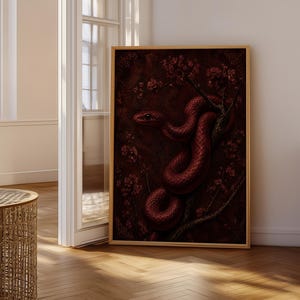 May include: A rectangular wall art print in a thin gold frame featuring a dark, deep red snake coiled around a branch with small, matching red blossoms. The artwork is set against a dark, textured background with a moody, monochromatic colour palette.