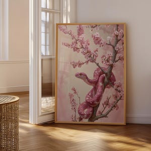 May include: A rectangular framed wall art print featuring a pink snake coiled around a flowering cherry blossom branch. The artwork displays soft pink and white floral petals against a muted pink background, encased in a thin gold frame.