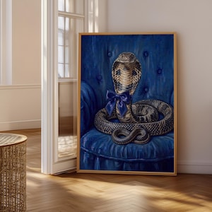 May include: A framed rectangular wall art print featuring a detailed painting of a cobra snake with a dark blue satin bow tied around its neck. The snake is coiled on a tufted, deep blue velvet armchair, rendered in a realistic artistic style.