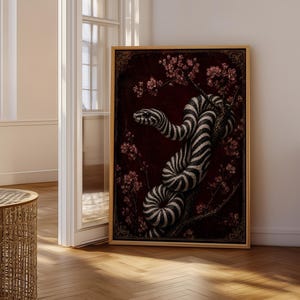 May include: A rectangular framed wall art print featuring a black and white striped snake coiled around a branch. The background is a deep dark red colour with small pink floral blossoms scattered throughout the composition in a vertical orientation.