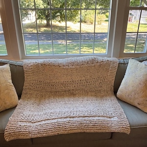 May include: A rectangular hand-knitted throw blanket in a light beige color with subtle dark brown flecks throughout. The blanket features a thick, textured garter stitch pattern with a distinct border framing the central section of the knit fabric.