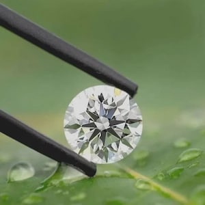 May include: A round, brilliant-cut, clear gemstone held by black metal tweezers. The stone features multiple triangular and kite-shaped facets that reflect light, creating a bright, sparkling appearance with sharp, geometric edges throughout.