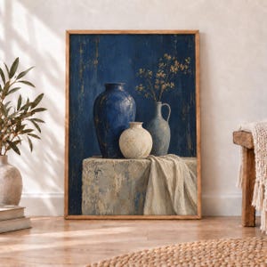 May include: A framed wall art print featuring a still life composition of three ceramic vessels on a stone block. A large dark blue vase, a smaller round cream vase, and a medium blue pitcher with delicate dried branches are set against a textured dark blue background with a draped cream fabric.