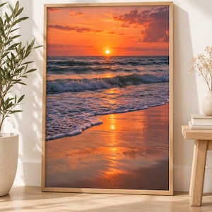 May include: A rectangular framed wall art print featuring a vibrant sunset over the ocean. The scene displays a bright orange sun low on the horizon, casting a golden reflection across the wet sand and gentle waves rolling onto the beach shore.