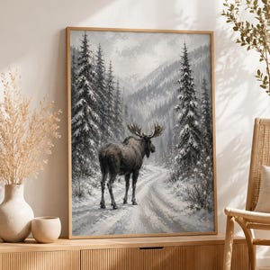May include: A framed rectangular wall art print featuring a dark brown moose standing on a snow-covered path. The scene depicts a winter forest landscape with tall evergreen trees dusted in white snow, set against a backdrop of misty mountains.