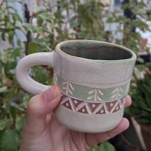May include: A ceramic mug with a cream-colored crackle glaze finish. The mug features a horizontal band decorated with carved green tree motifs and a lower band of carved reddish-brown triangles with central dots. It has a thick, curved handle.