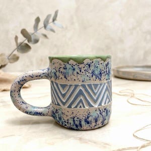 May include: A ceramic mug featuring a light green rim and a body decorated with blue and white speckled patterns. A central horizontal band displays a carved white chevron design. The mug has a curved handle and a speckled finish throughout.