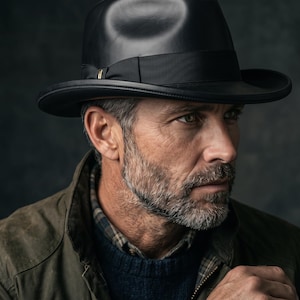 Pode incluir: Um chapéu fedora preto com aba larga e uma fita de gorgorão preta com um pequeno detalhe metálico. O chapéu é feito de um material liso e estruturado, usado sobre um suéter azul-marinho escuro e uma jaqueta verde-oliva.