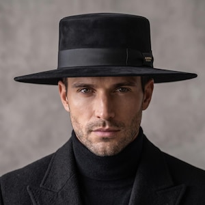 Puede incluir: Un sombrero negro de copa plana con ala ancha y una cinta de satén negra alrededor de la corona. El sombrero presenta un pequeño detalle metálico rectangular de tono dorado en el lateral de la cinta. Se lleva con cuello alto y abrigo.