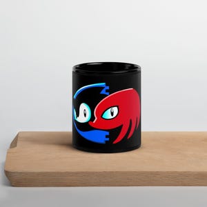 May include: A black ceramic mug featuring a graphic design of two stylised character profiles facing each other in a yin-yang formation. One side is a blue and white silhouette of a hedgehog, while the other is a red and white silhouette of an echidna.