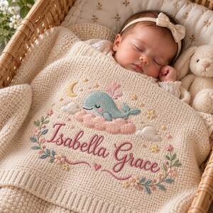 Puede incluir: Una manta de punto para bebé en color crema con un diseño bordado de una ballena azul sobre una nube rosa, una luna creciente amarilla, estrellas y el nombre Isabella Grace en letra cursiva rosa, rodeado por un borde floral.