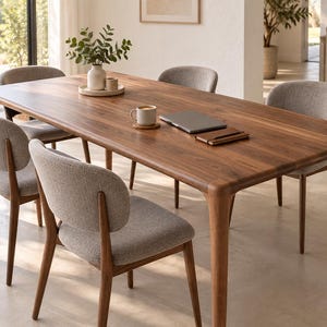 May include: A rectangular wooden dining table with rounded corners and four tapered legs, accompanied by six chairs featuring light grey fabric upholstery and wooden frames. A ceramic vase with greenery, a coffee mug, and a laptop sit on the table.