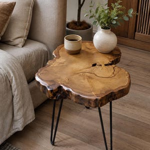 May include: A side table featuring a natural live-edge wood slab top with visible grain and a central crack, supported by three slender black metal hairpin legs. The surface holds a small ceramic cup and a round white vase with green foliage.