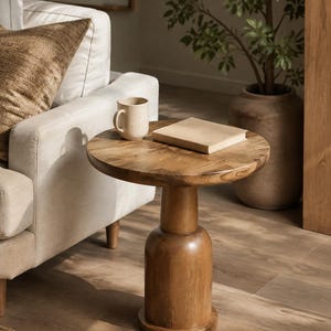 May include: A round wooden side table with a thick, pedestal-style base. The table features a smooth, circular top surface with a visible wood grain pattern. The base is cylindrical and flares slightly toward the top, resting on a flat circular foundation.